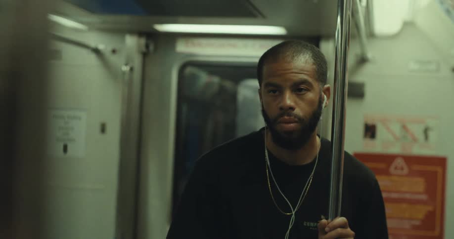 A sleepylooking man with a beard and earphones holds onto a metal pole for balance as he stands inside a shaking subway car looking around his surroundings