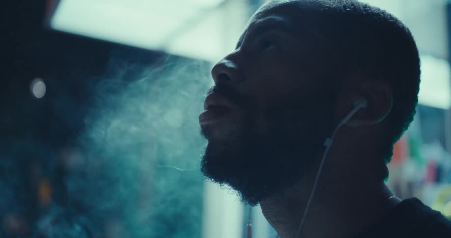 In a moody closeup at a brightly lit bus stop a man with earphones lights a handrolled cigarette takes a deep puff and exhales a thick cloud of smoke as he tilts his head back