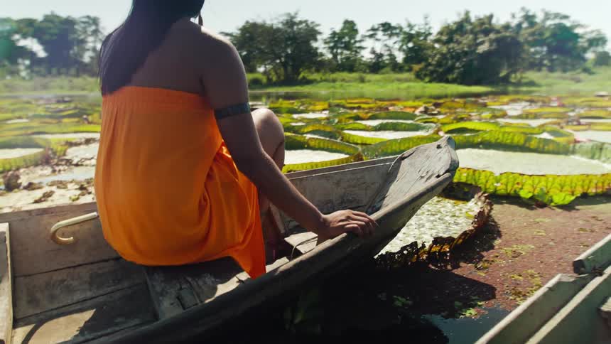 From a tracking boat the camera follows a woman in a bright orange wrap as she sits in a wooden canoe and glides through a sunlit lake covered in giant round lily pads