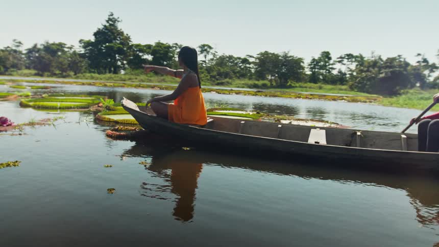 A woman in a red shirt paddles a wooden canoe on a sunlit lake as another woman in an orange dress sits in the front and points forward navigating past giant floating lily pads