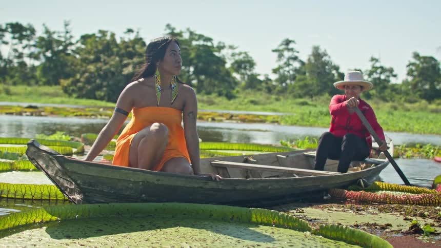 A woman in an orange dress sits at the bow of a wooden boat while another woman paddles through a lilycovered waterway