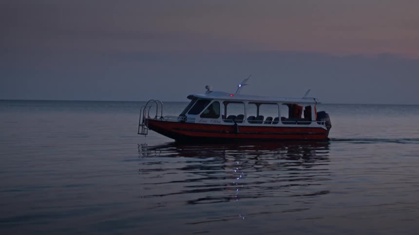A small passenger boat glides across calm water at dusk its lights reflecting on the surface