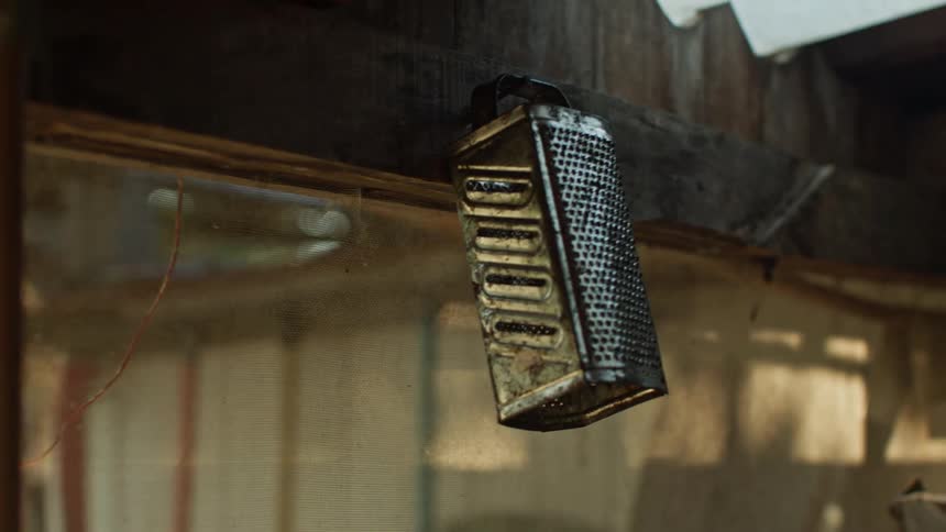 A close view shows an old tarnished metal cheese grater hanging from a wooden beam