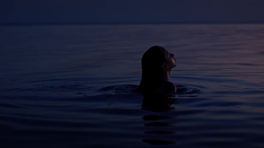 A person emerges from the dark water their head and shoulders visible against the twilight sky their silhouette is reflected on the rippling surface of the water as they look upwards