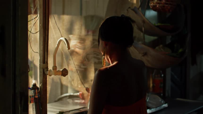 A person in an orange garment is seen from behind preparing something in a kitchen sink area the space is illuminated by a warm light coming from the window highlighting the person and the cluttered kitchen environment