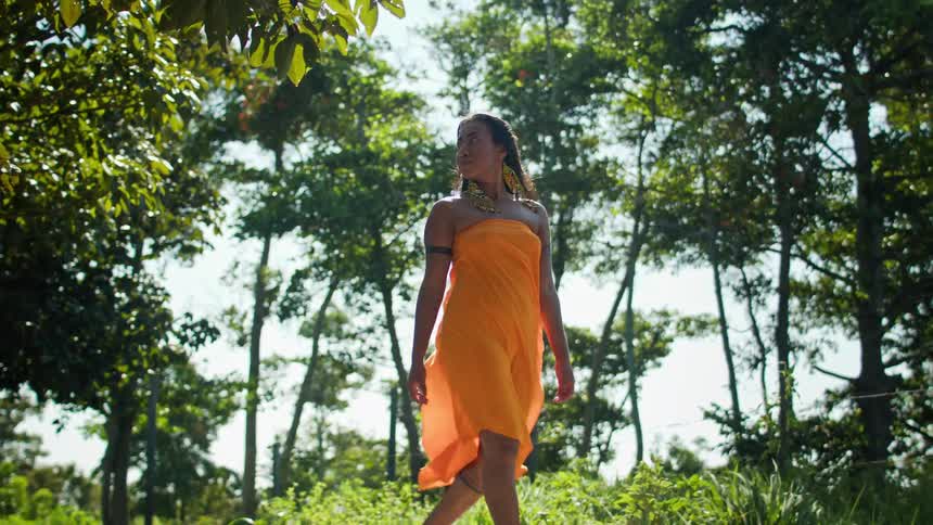 A woman in an orange dress walks through a sunlit grove full of trees