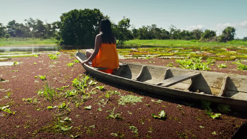 A wide shot from the side shows a person in an orange wraparound dress sitting in the front of a small wooden boat the boat is in a shallow river filled with water plants with another person joining in the back to paddle