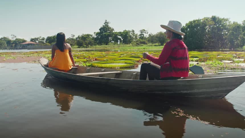 Two women in a wooden boat rowing near aquatic plants
