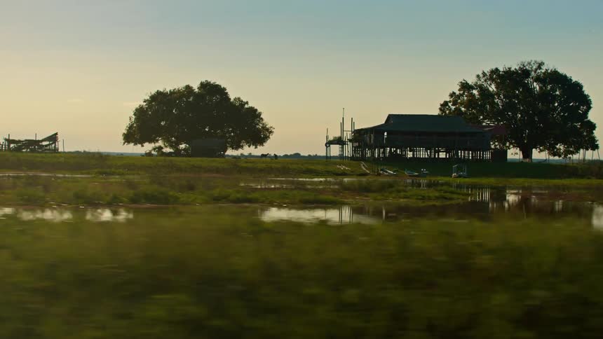 A tracking shot glides past a calm marsh at sunset revealing the dark silhouette of a stilt house and large trees which are perfectly reflected in the still water below