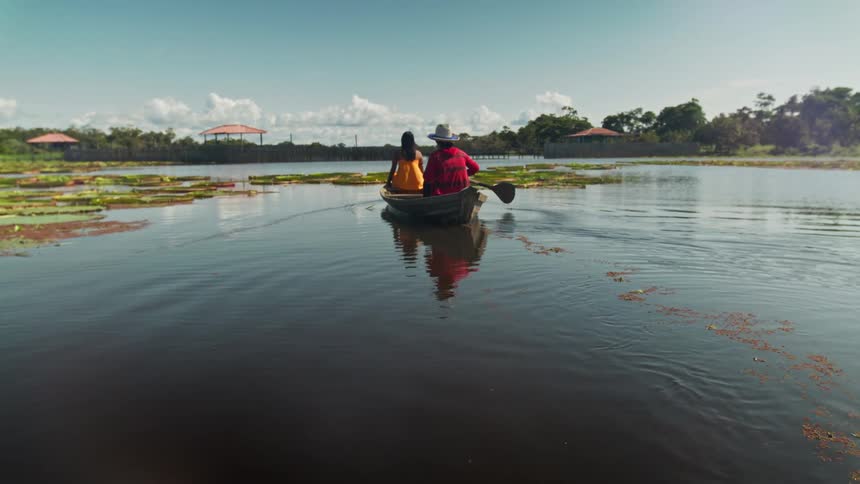 Two people are seen from behind paddling a small boat across a calm body of water filled with lily pads under a clear blue sky