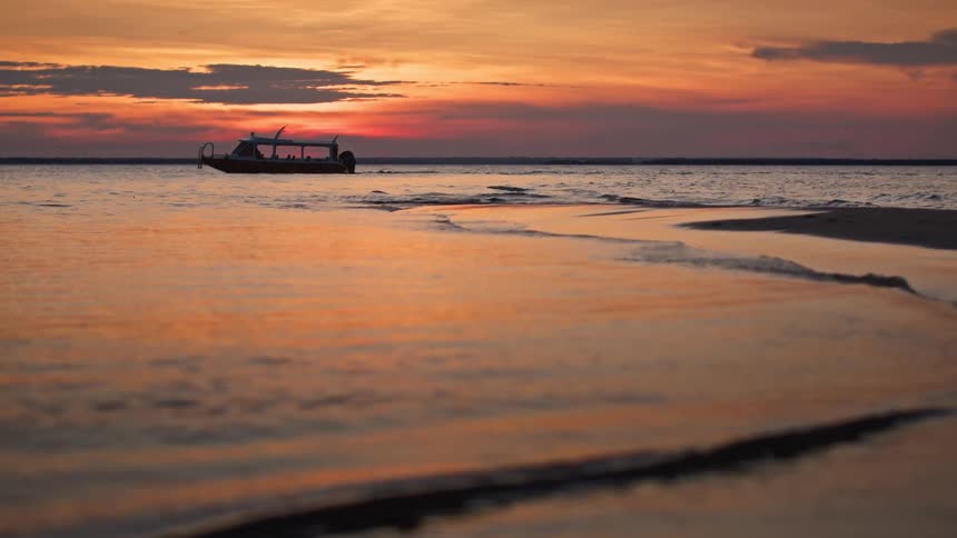 A small boat floats on the water near a sandy shore during a vibrant sunset with orange and purple hues