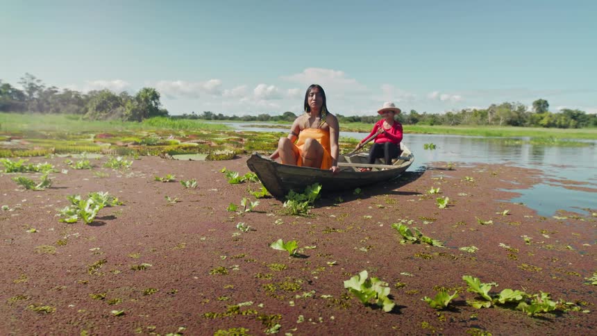 Two women in a wooden boat navigate through an area covered with aquatic vegetation