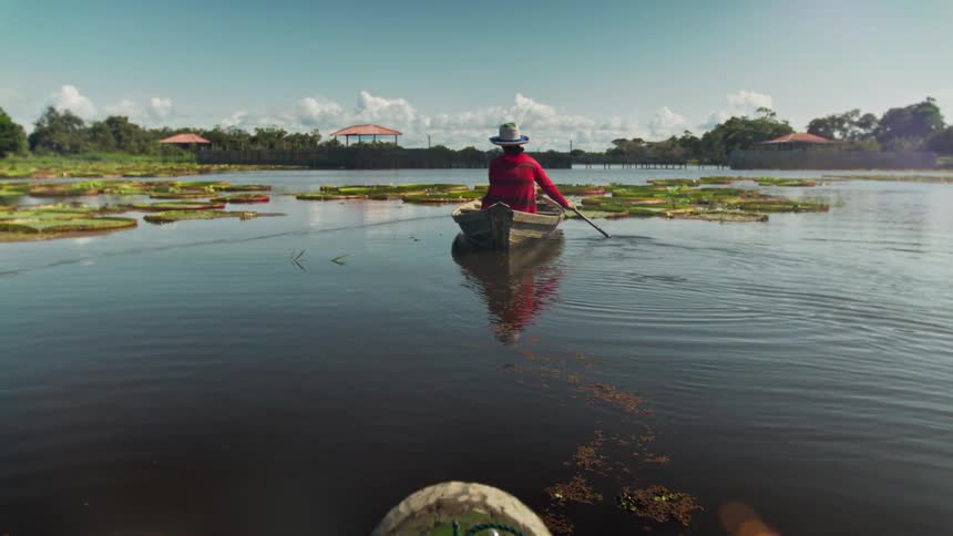 Two women row a wooden boat surrounded by aquatic plants