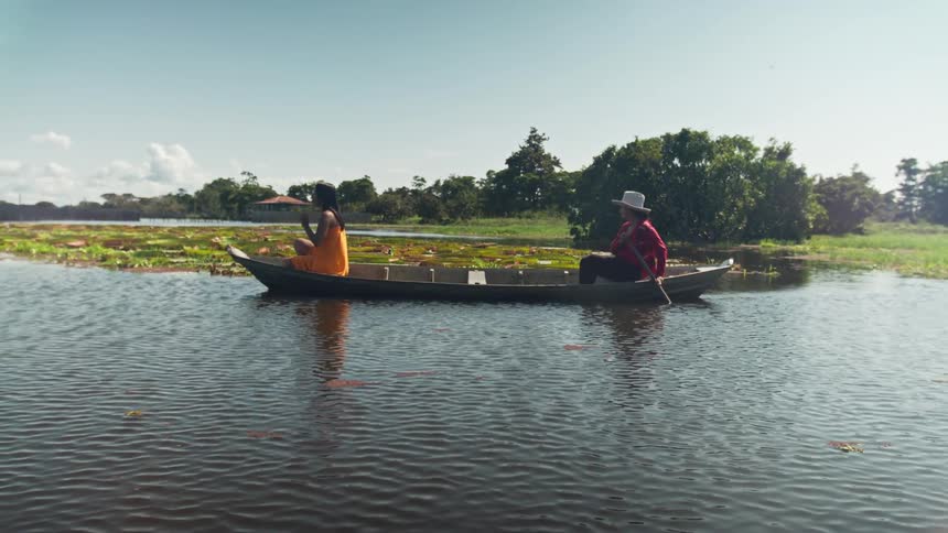 Two women ride a canoe on a river at midday one sitting at the front and the other at the back wearing sun hats and paddling the canoe forward