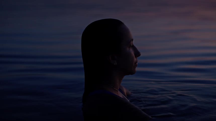A person floats in dark calm water at dusk gazing toward the fading light on the horizon