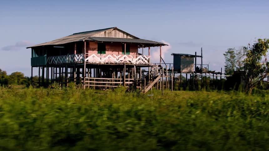 A stilted wooden house with a patterned railing stands above grassy wetlands beside a calm waterway under a clear sky