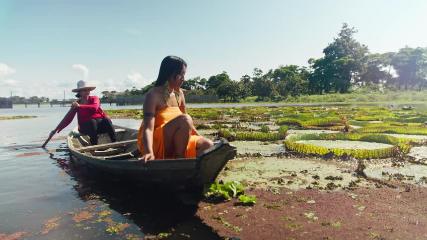 Woman in a wooden boat looks at aquatic plants while another rows