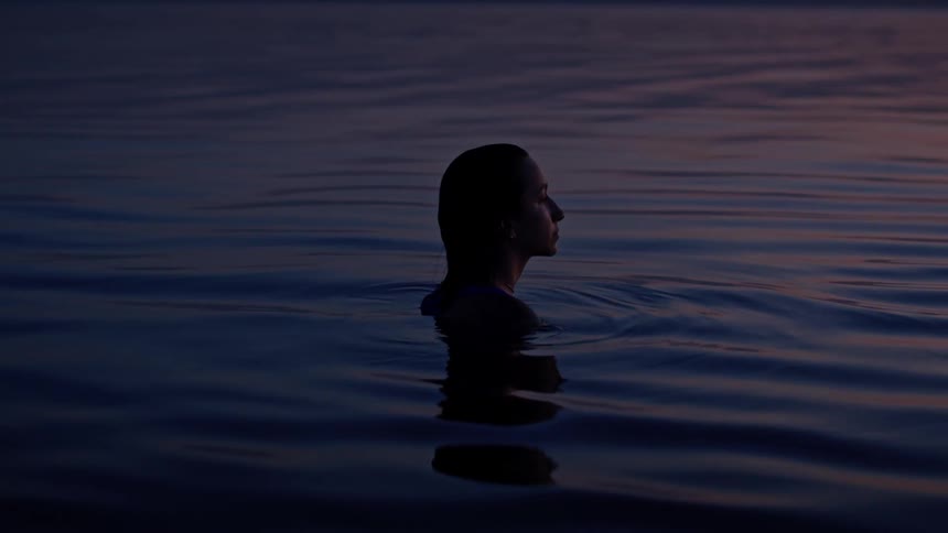A woman with long dark hair is standing in a lake at sunset with her shoulders and head visible above the water as her silhouette is visible against the pink and purple sky while she looks to her right