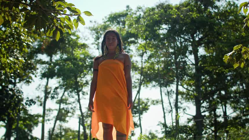 A woman with dark hair wearing a loose orange dress is standing in a lush forest and looking directly at the camera with a neutral expression on her face