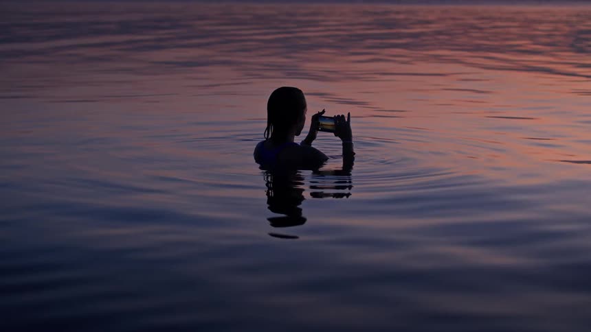 A woman with her hair tied back is standing in the middle of a lake at sunset with the water coming up to her chest as she holds a smartphone in both hands to take a video of the view in front of her