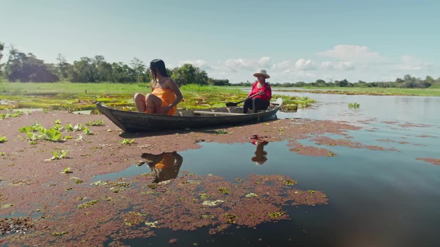 Two people sit in a small boat surrounded by reddish brown aquatic plants on a sunny day