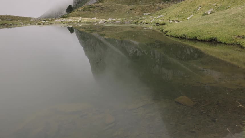A calm body of water reflects a mountain peak and the misty sky bordered by grassy slopes