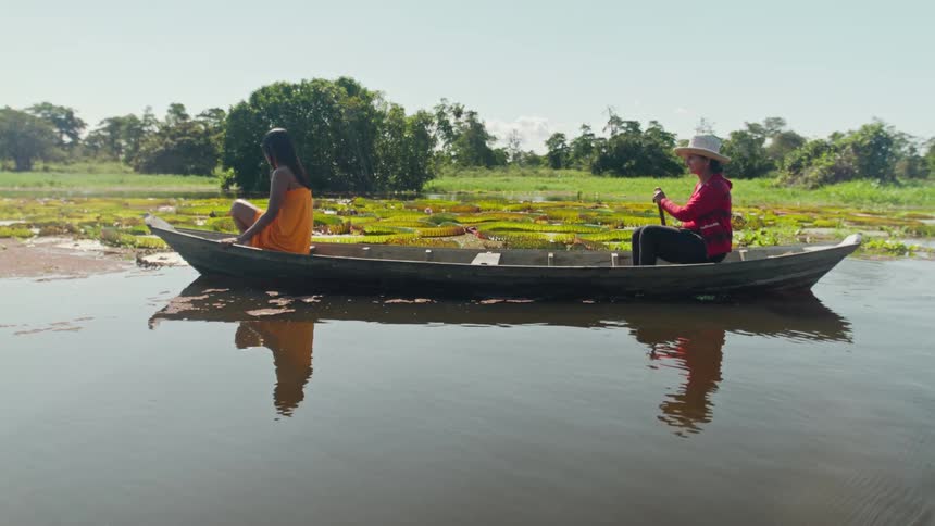 Two women one wearing a hat and a red long sleeve shirt and the other wearing an orange dress are sitting in a small boat on a lake filled with giant lily pads as they paddle and talk together