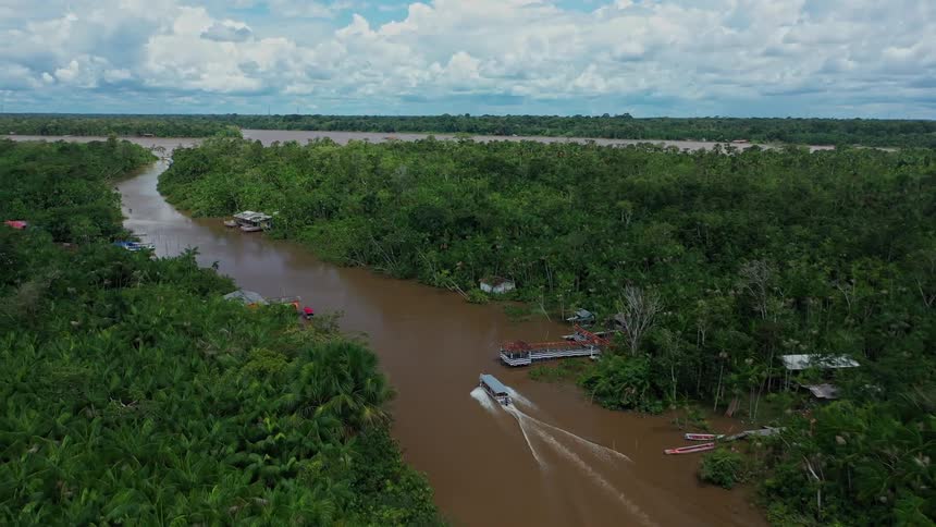 A brown river winds through a dense green forest with a boat leaving a white wake and a small settlement of houses and docked boats visible along the banks