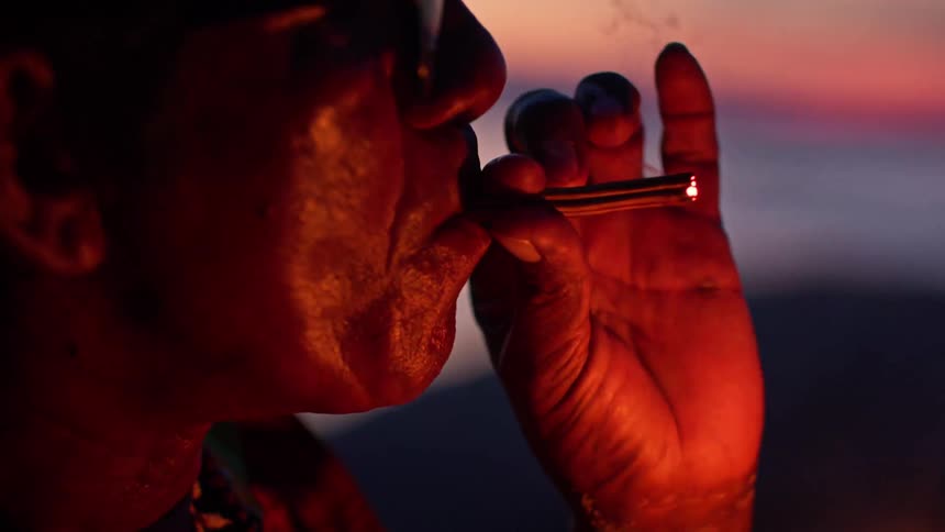A close view shows the silhouette of a person with glasses as they hold a match up to a cigar in their mouth and a sunset is visible over the ocean in the background