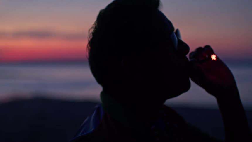 A man wearing glasses smokes by the water at sunset the glowing ember of his cigarette standing out against his silhouette