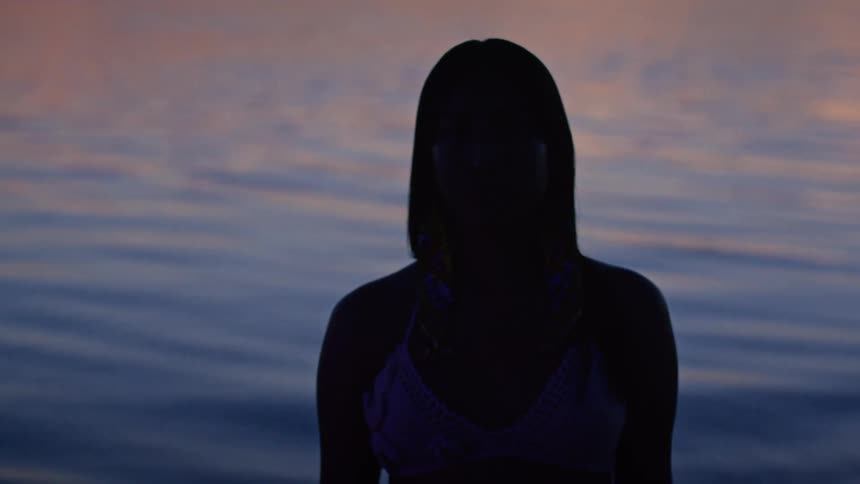 A woman with long dark hair is standing on a beach at sunset with her back to the camera as the camera slowly pans around to show her face in silhouette against the water