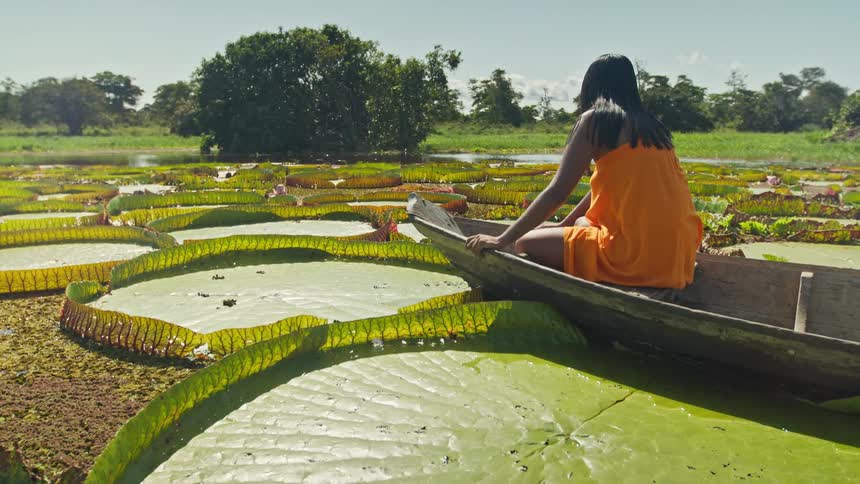 A woman is in a wooden boat and wearing an orange dress and she is touching the surface of a water lily