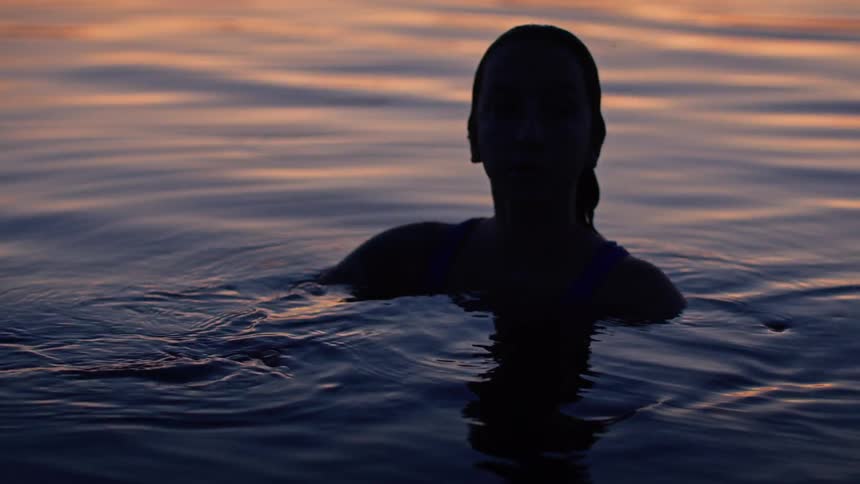 A woman with long hair is standing in a lake at sunset with her shoulders and head visible above the water as her silhouette is visible against the fading light on the horizon while she looks to her right