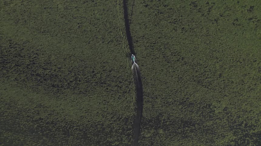 An aerial highangle shot captures a person on a small motorized scooter traveling along a narrow dirt path the path cuts through a vast green field and the persons bright clothing stands out against the dark landscape