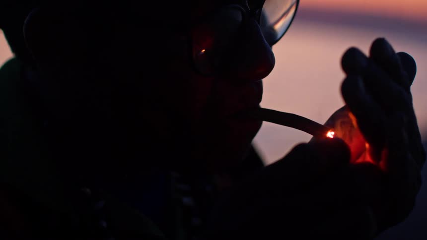 A close view shows the silhouette of a person with glasses as they hold a lighter up to a cigar in their mouth and a sunset is visible over the ocean in the background