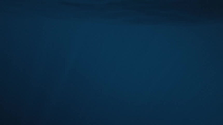 The camera is submerged underwater showing a deep blue expanse it then breaks the surface revealing a closeup shot of sunlit waves crashing
