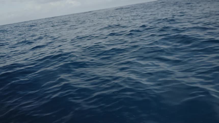 The video shows a view from a moving boat capturing the vast deep blue ocean under a partially cloudy sky the camera pans down to show the turbulent foamy waves created by the boats movement