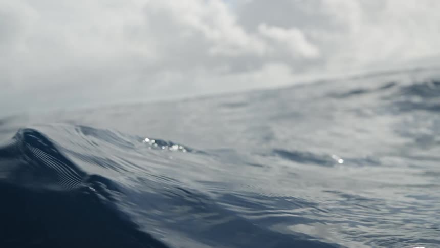 A low angle view from inside a small boat shows the waves of the dark blue ocean with a cloudy sky above as the boat moves up and down on the water and the waves crash into the side of the camera