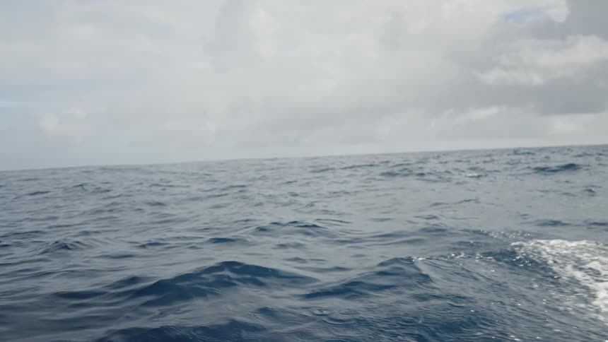 The video shows a view from a moving boat with waves of water being pushed away from the side the vast blue ocean stretches out to the horizon under a cloudy sky