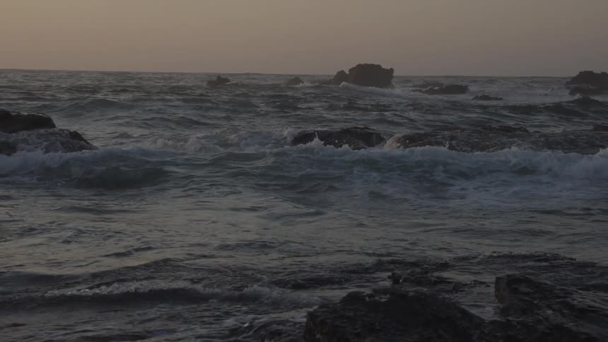 Waves crash over dark rocks in the ocean at sunset