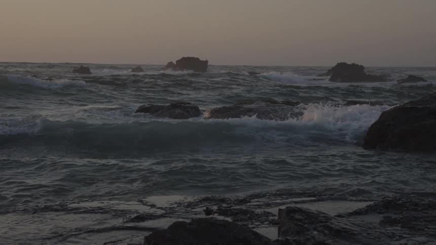 A long view shows a dark ocean with many large waves crashing on a rocky coastline with several dark rock formations rising out of the water under a cloudy sunset sky