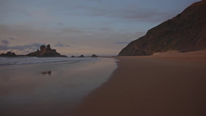 Waves roll onto a beach beside a rocky cliff at sunset