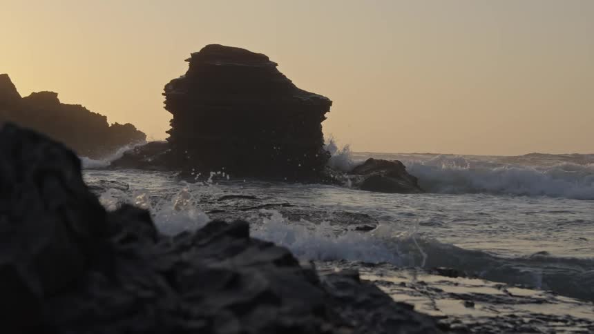 Waves crash against jagged rock formations along the shore at sunset with a large sea stack silhouetted against the golden sky