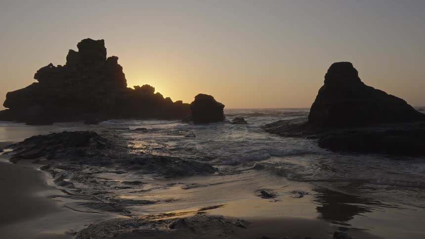 Waves churn onto the beach passing coastal rock formations silhouetted against the sunlight on the red tinted horizon