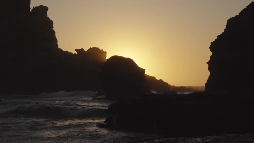 The sun sets behind silhouetted rocks on a coastline casting a golden glow over the water waves crash against the rocky shore as the light fades creating a dramatic and serene seascape