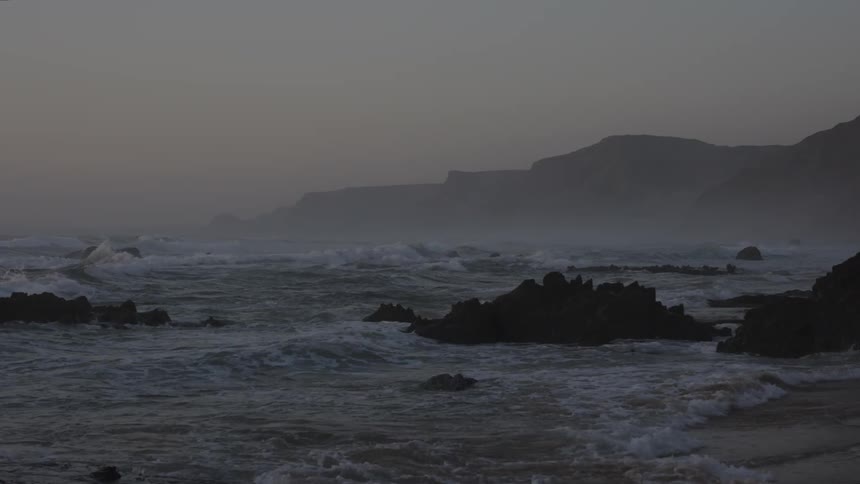 The sea water splashes against a rocky coast at dusk