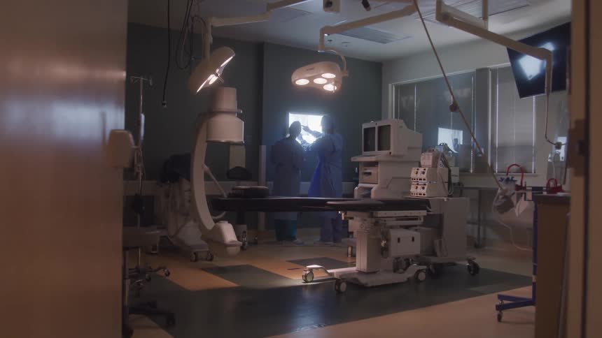 A medical team in blue scrubs stands in a brightly lit operating room discussing a patients scans the room is filled with various medical equipment including an operating table monitors and an xray light box