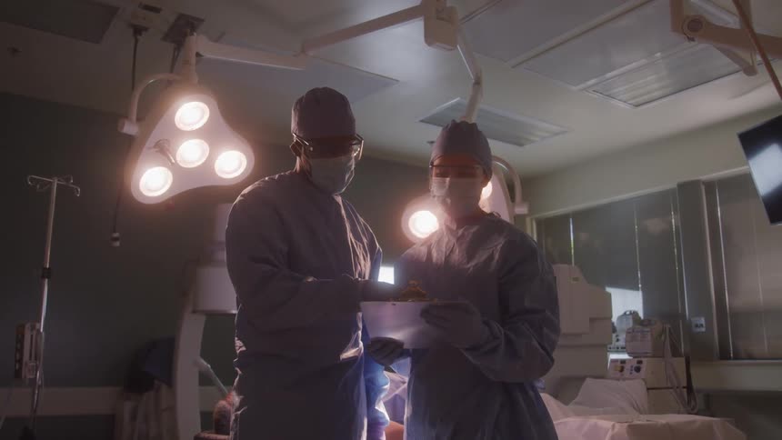 Two surgeons wearing masks and sterile gowns work in a brightly lit operating room