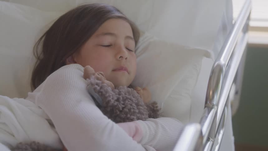 Anxious little girl laying in a hospital bed hugging her teddy bear