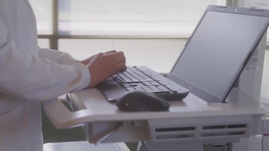 A medical professional in a white coat is standing and typing on a laptop the laptop is on a mobile medical cart suggesting they are working in a hospital or clinic setting
